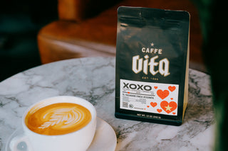 12oz bag of XOXO front, on marble table next to latte in a ceramic cup with saucer
