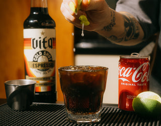 Hand juicing a lime over a glass of Cold Shot Coke next to a bottle of Vita Cold Shot, shot glass, can of Coca-Cola, and lime.