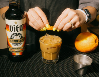 Hands zesting an orange peel above a glass filled with an iced Valencia next to a bottle of Vita Cold Shot, shot glass, and orange.