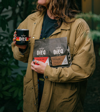 Person holding camp cup with Caffe Vita sunset logo, 12oz bag of Caffe Del Sol, and 12oz bag of Theo Blend in front of green bushes