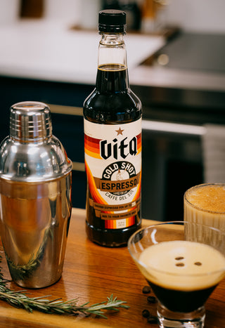 Vita Cold Shot on bar with various cocktails, cocktail shaker, and sprig of rosemary.