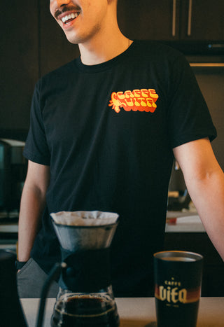 Person wearing black t-shirt with yellow and orange scorpion design and Caffe Vita logo, standing in a kitchen with a pourover and Vita tumbler