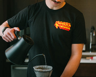 Person wearing black t-shirt with yellow and orange scorpion design and Caffe Vita logo, standing in a kitchen with a pourover and Vita tumbler