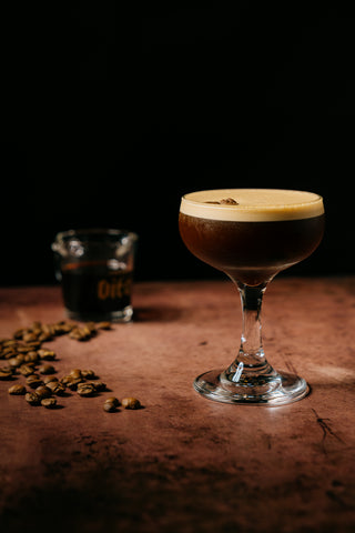 Espresso martini in coup on counter with coffee beans and shot glass of espresso