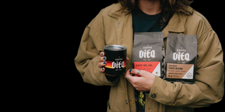 Person holding camp cup with Caffe Vita sunset logo, 12oz bag of Caffe Del Sol, and 12oz bag of Theo Blend on black background