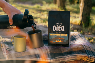 12oz bag of Campfire blend on blanket with cups being filled with coffee as fire burns in foreground. 