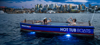 Two people relaxing in a hot tub boat on water with a city skyline in the background.