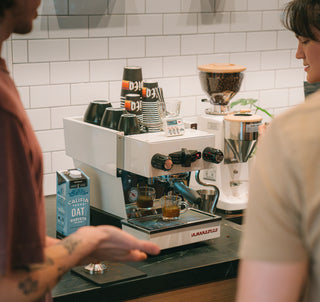 People standing in front of a mini espresso machine pulling espresso into shot glass with Vita branded to go cups and ceramic mugs on top 